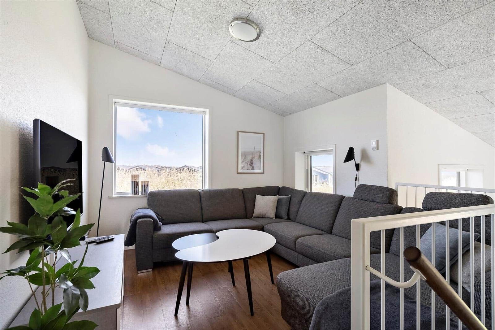 Wohnzimmer mit grauem Sofa, runden Tisch und TV. Fenster mit Blick auf Landschaft.