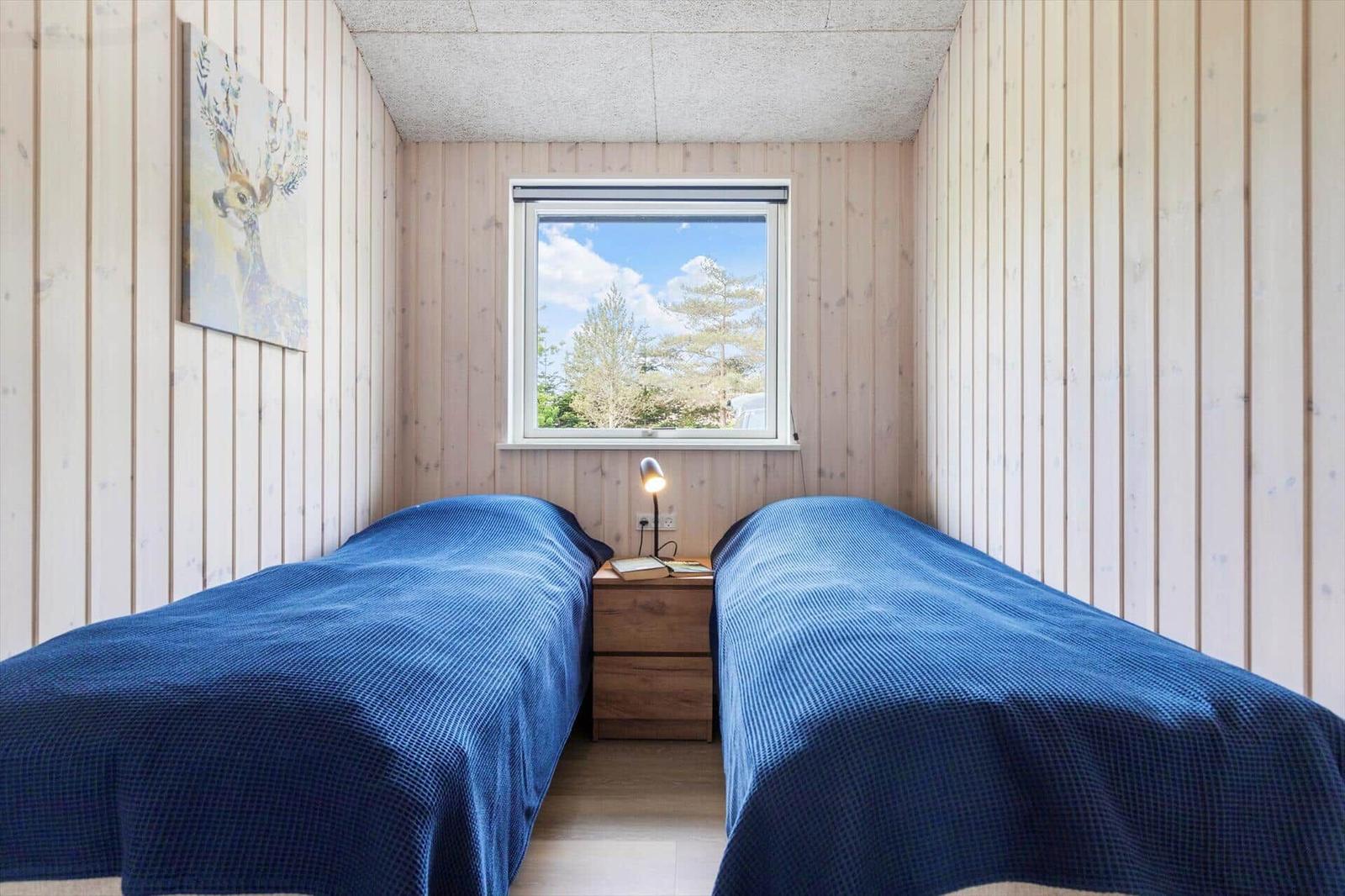 Two beds with blue covers, wooden walls, window with view of trees.