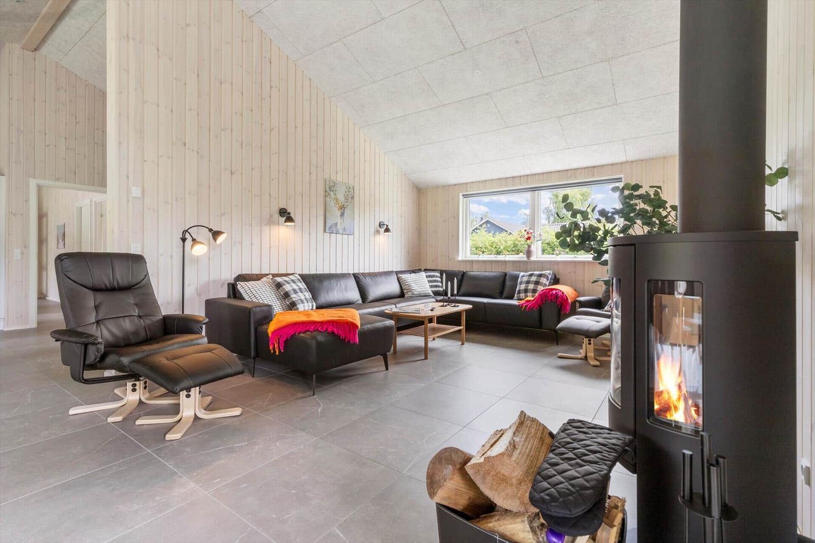 Living room with wooden walls, sofa, chair, and wood stove. Window with view outside.