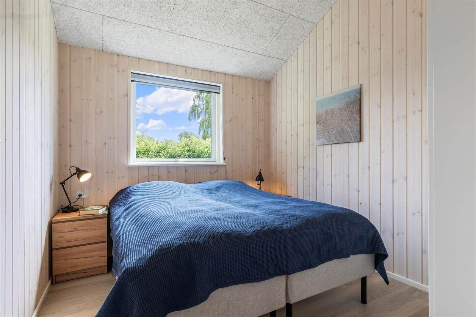 Bedroom with wooden walls, bed with blue blanket, and window with view of trees.