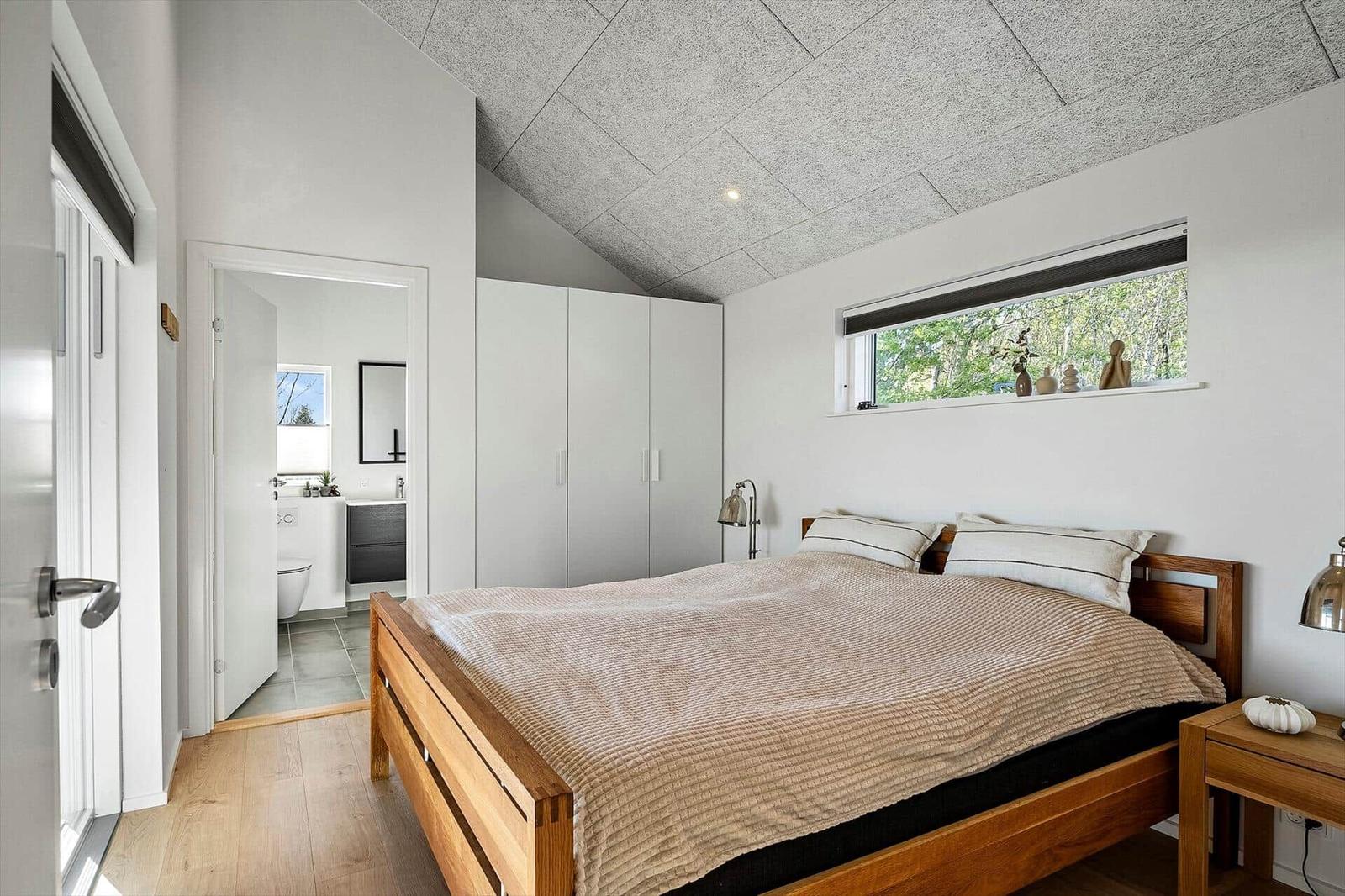 Schlafzimmer mit Holzbett, Fenster und angrenzender Toilette.