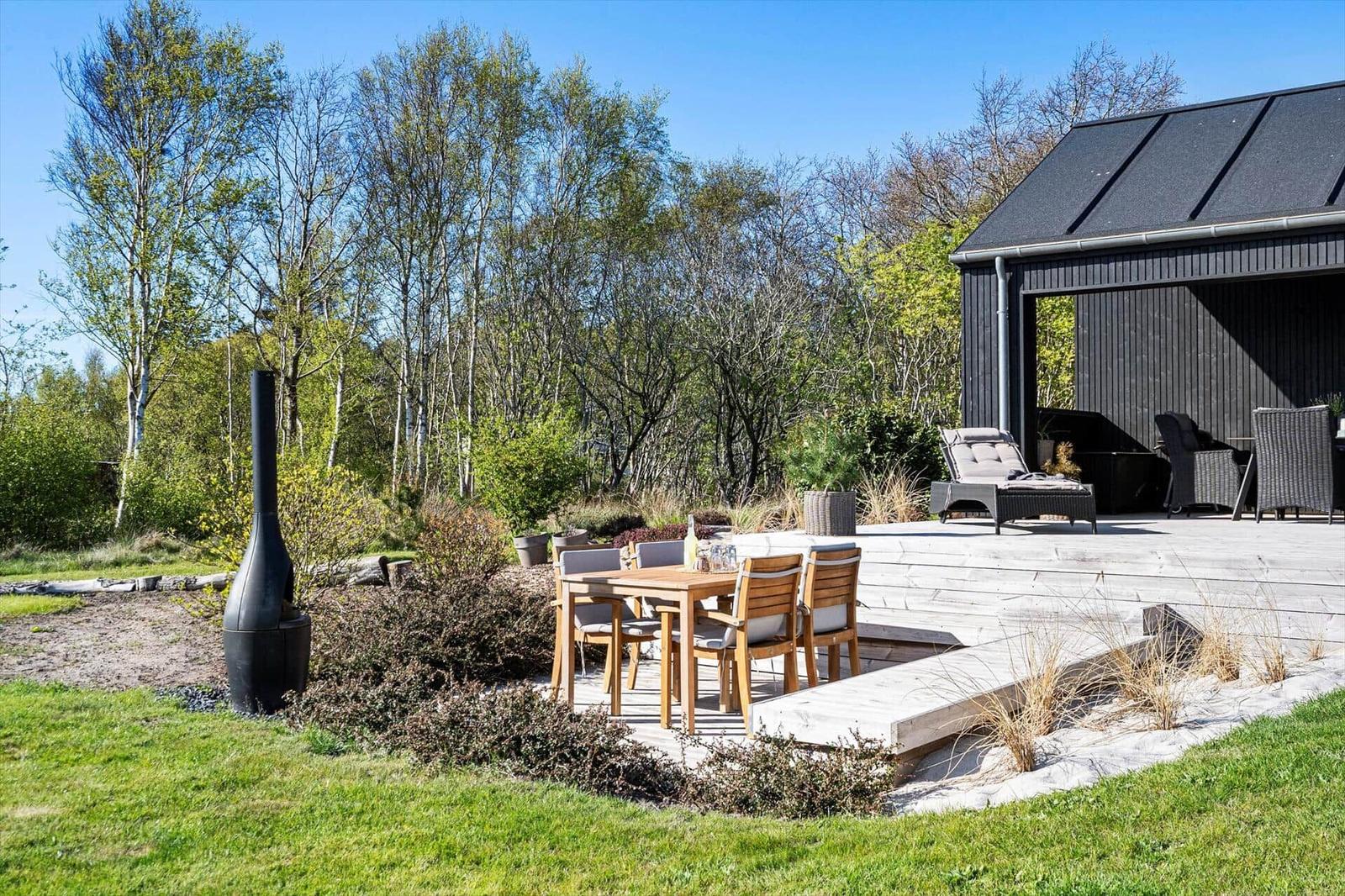 Terrasse mit Holztisch, Stühlen und Liege. Schwarzer Ofen und Gartenhaus im Hintergrund.