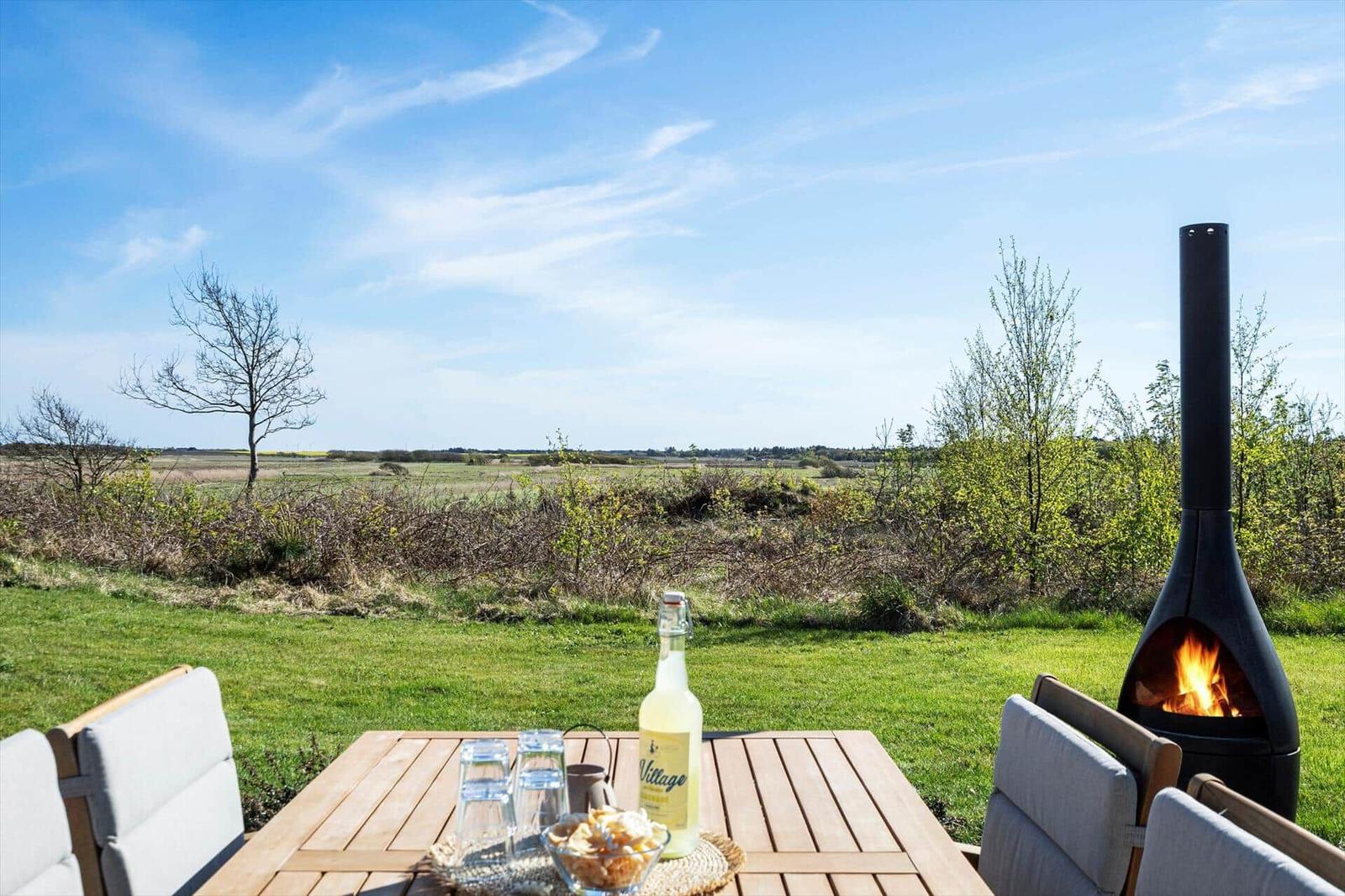Außenbereich mit Tisch, Stühlen und Feuerkamin. Blick auf grünes Feld und Himmel.