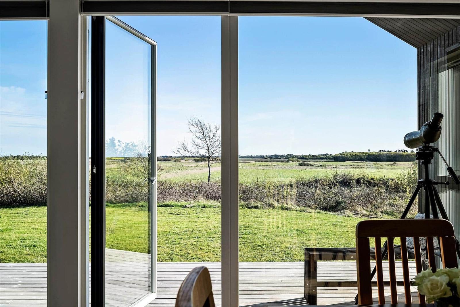 Große Fenster bieten Blick auf grünes Feld und klare Himmel. Holzterrasse mit Stuhl und Fernglas.