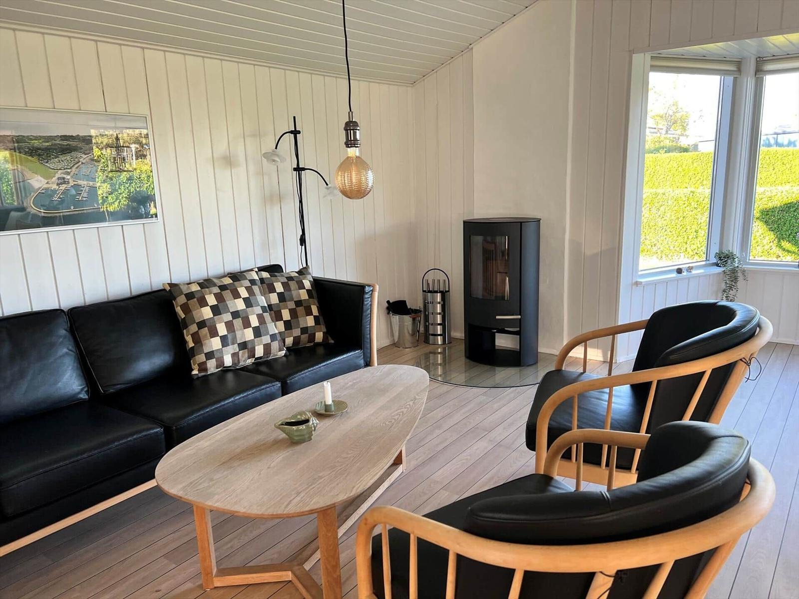 Wohnzimmer mit Sofa, zwei Stühlen, Tisch und Kaminofen. Fenster mit Blick auf Grünfläche.