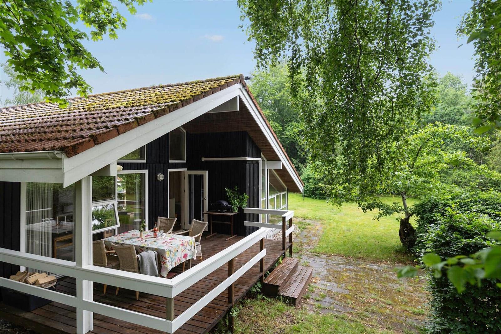 House with terrace, table and chairs, surrounded by greenery and trees.