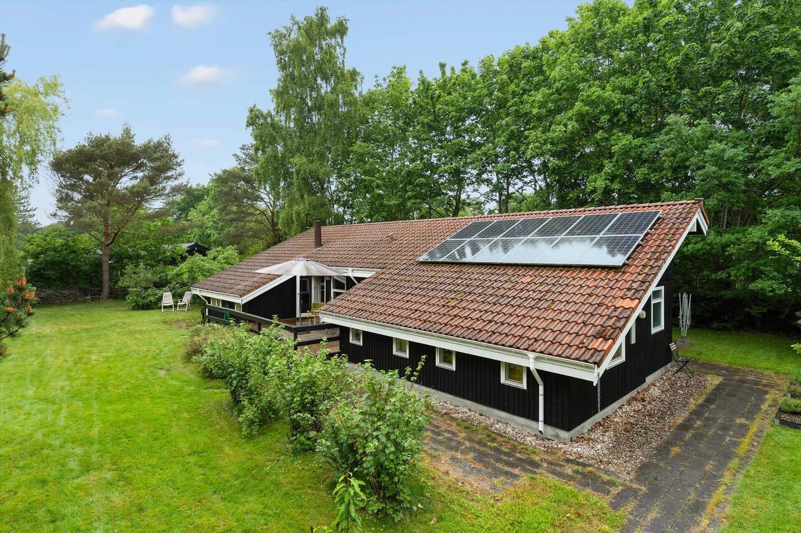 Black house with solar panels, terrace, and green garden. Surrounded by trees.