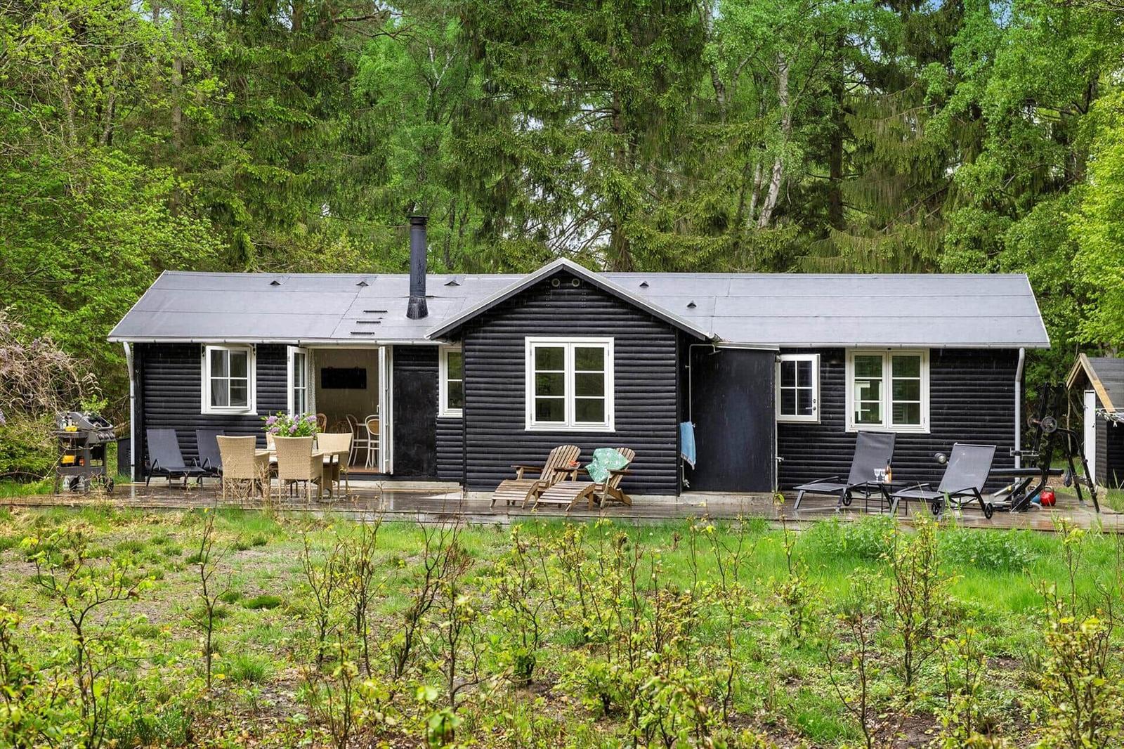Schwarze Holzhütte mit Terrasse, Garten und Grillplatz im Wald.