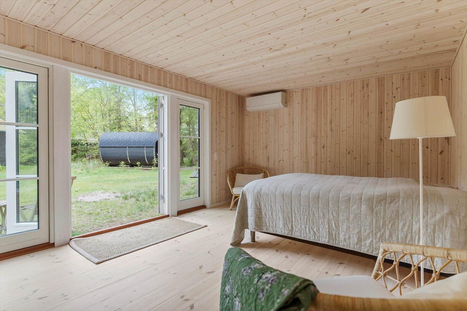 Schlafzimmer mit Holzwand, Bett, Stehlampe und Terrassentür zur Grünfläche.