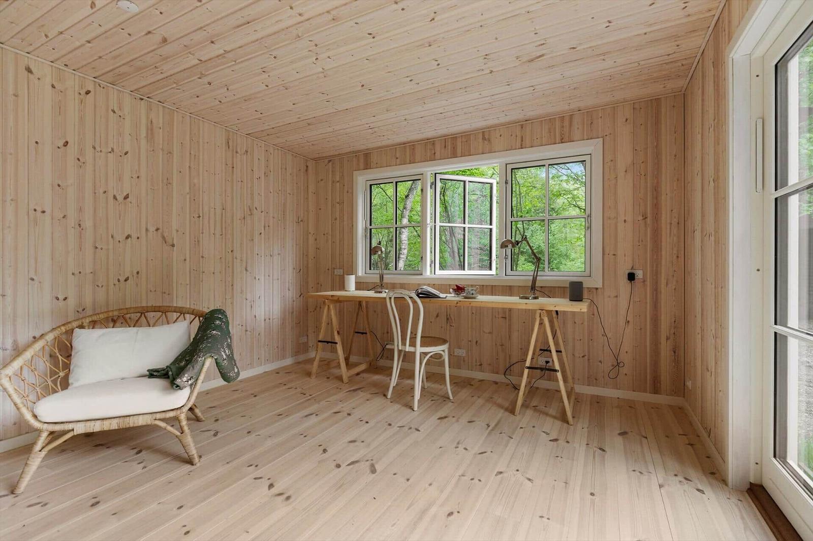 Raum mit Holzwand, Tisch, Stuhl und Sitzmöbel vor Fenster mit Blick auf Wald.