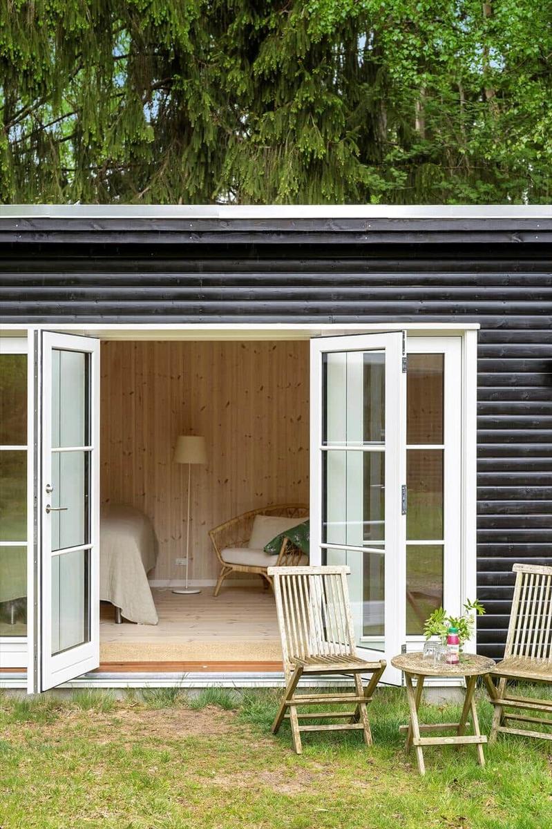 Schlafzimmer mit Bett, Stuhl und Stehlampe. Außen: Holzstühle und Tisch vor einem schwarzen Holzhaus.