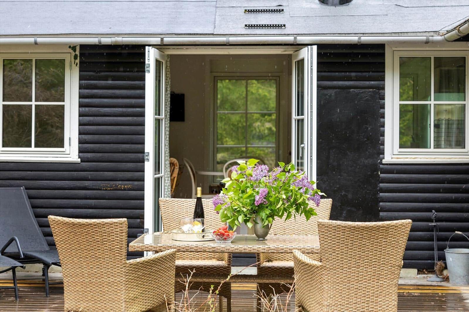 Terrasse mit Esstisch, Stühlen und Blumen vor schwarzem Holzhaus mit offenen Türen.