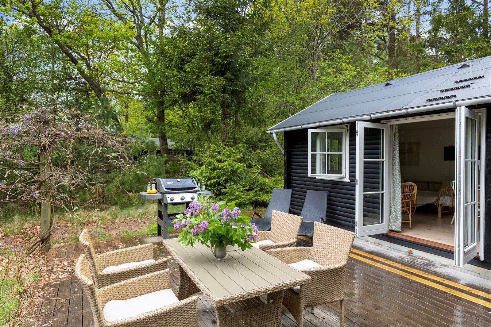 Terrasse mit Grill, Tisch und Stühlen. Im Hintergrund Wald und Ferienhaus mit offener Tür.