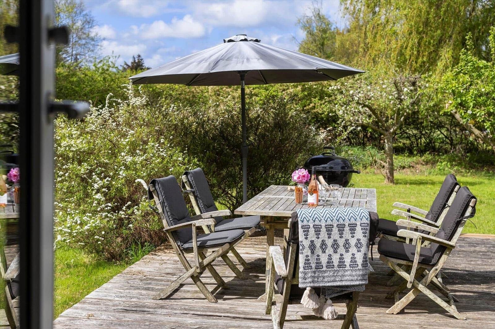 Deck with table, chairs, umbrella, and grill. Background: green garden with shrubs.