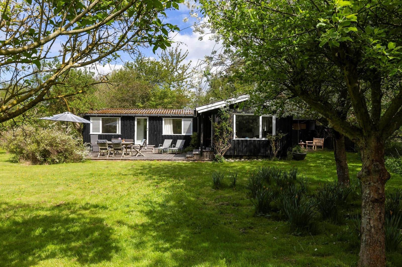 A house with a terrace, garden, and trees. There is a table with chairs and an umbrella.
