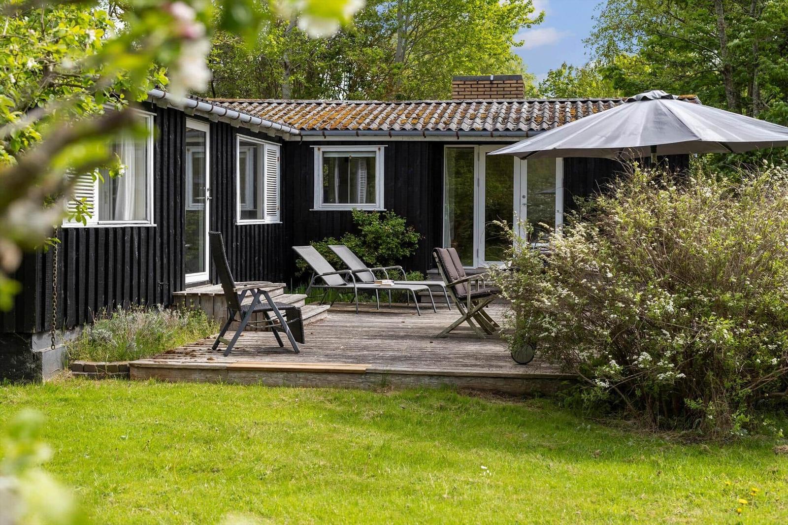Black wooden house with terrace, sun loungers, and umbrella. Surrounded by greenery.