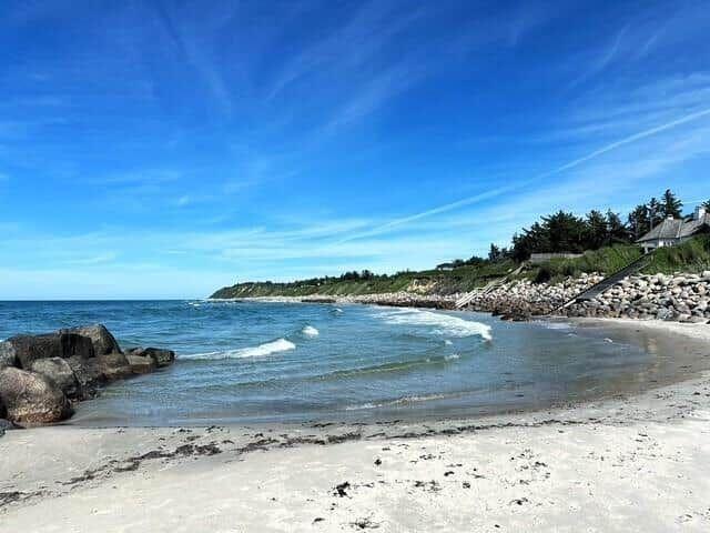 Beach with rocks and waves, blue sky, coastal road with trees and stones.