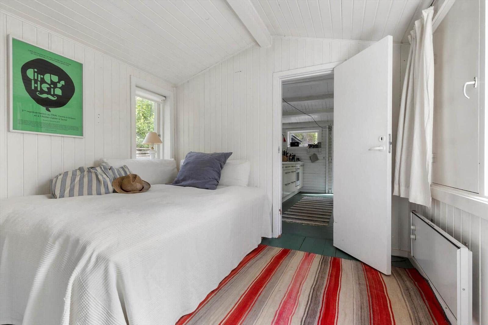 Bedroom with white bed, striped rug, and view into the kitchen.