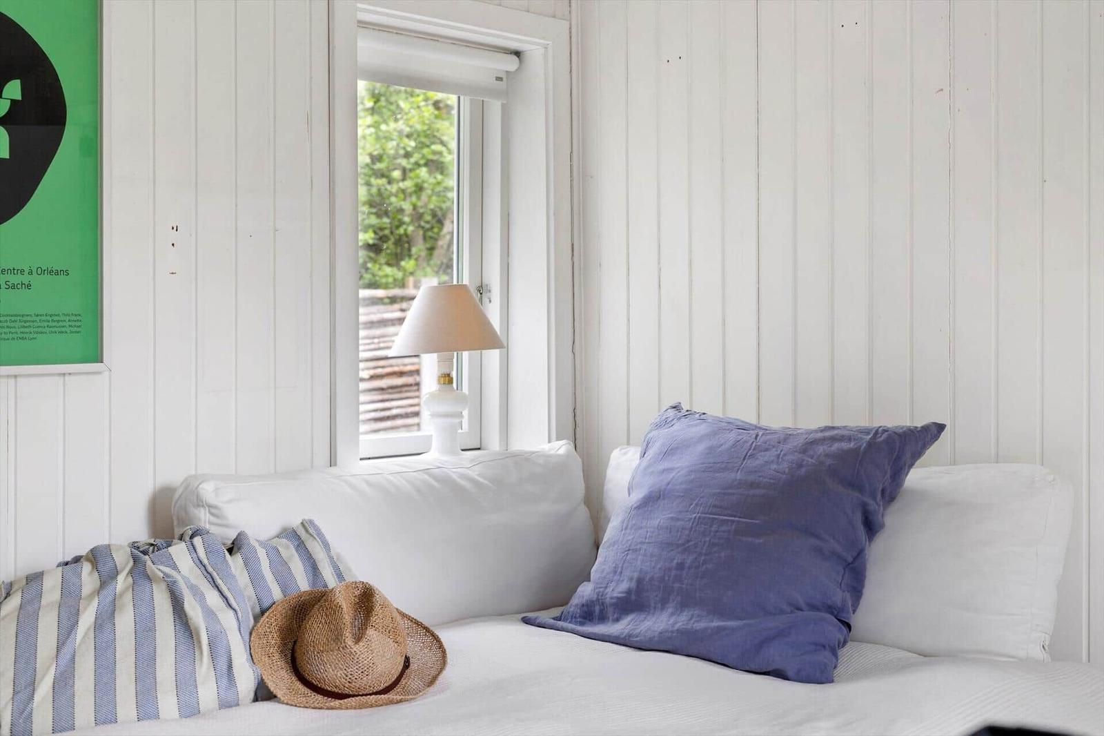 A bed with pillows and a hat by a window with a view of green.