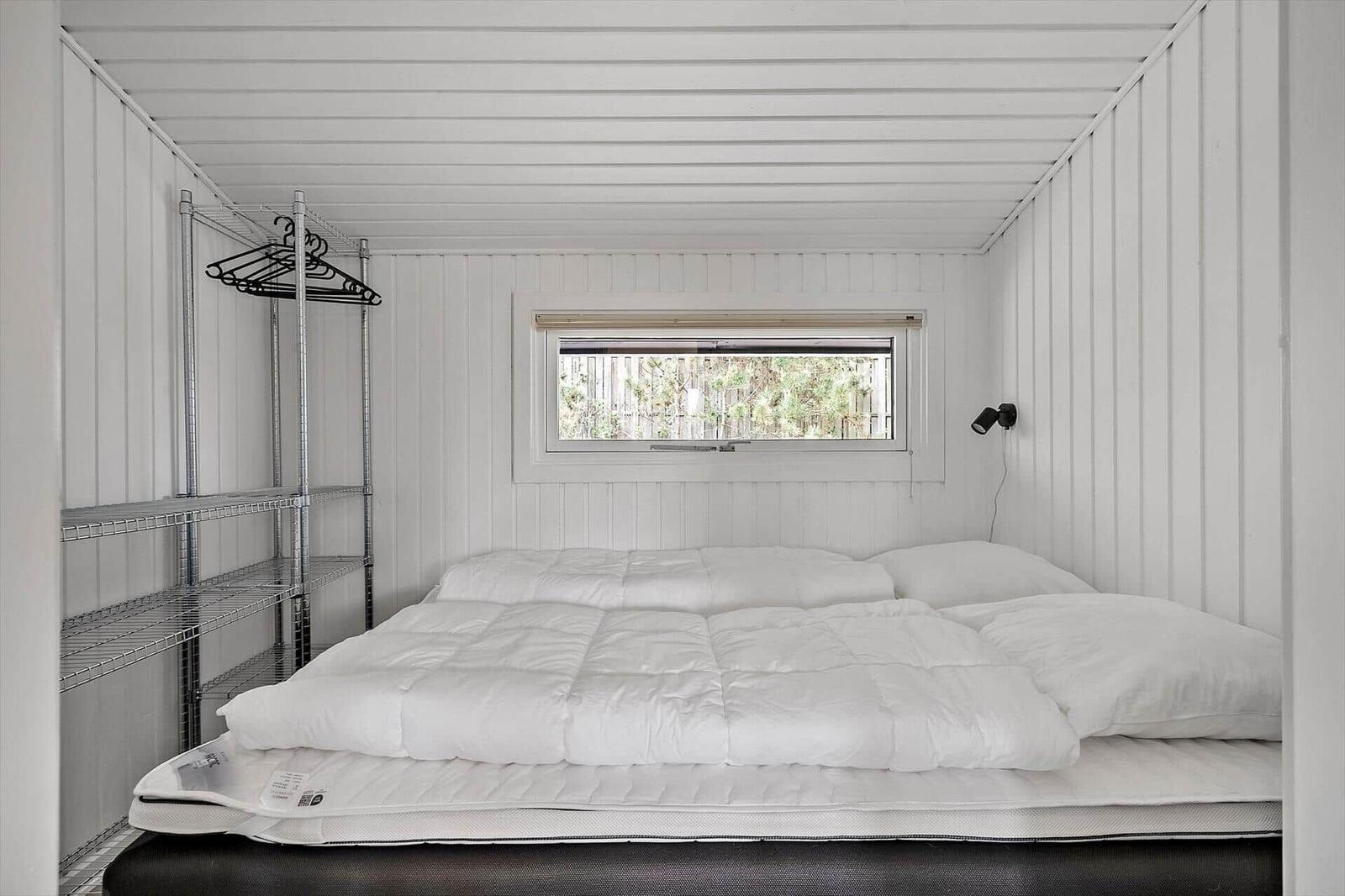 Schlafzimmer mit Matratze, Bettwäsche und Fenster. An der Wand steht ein Metallregal.