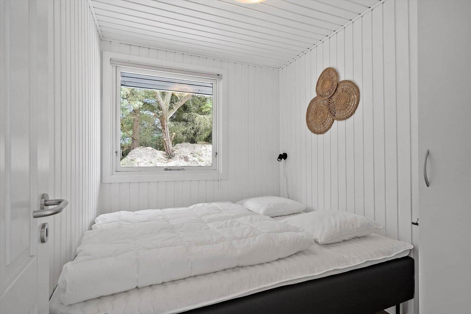 Schlafzimmer mit weißer Wandverkleidung, Bett und Fensterblick auf Bäume.