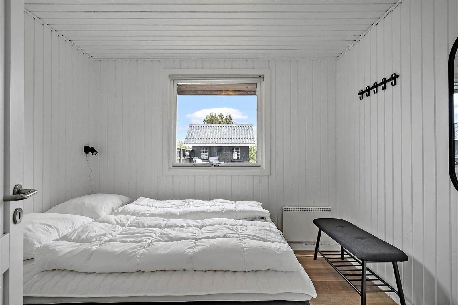 Schlafzimmer mit weißem Bett, Fenster und Bank. Wandverkleidung und Decke weiß.