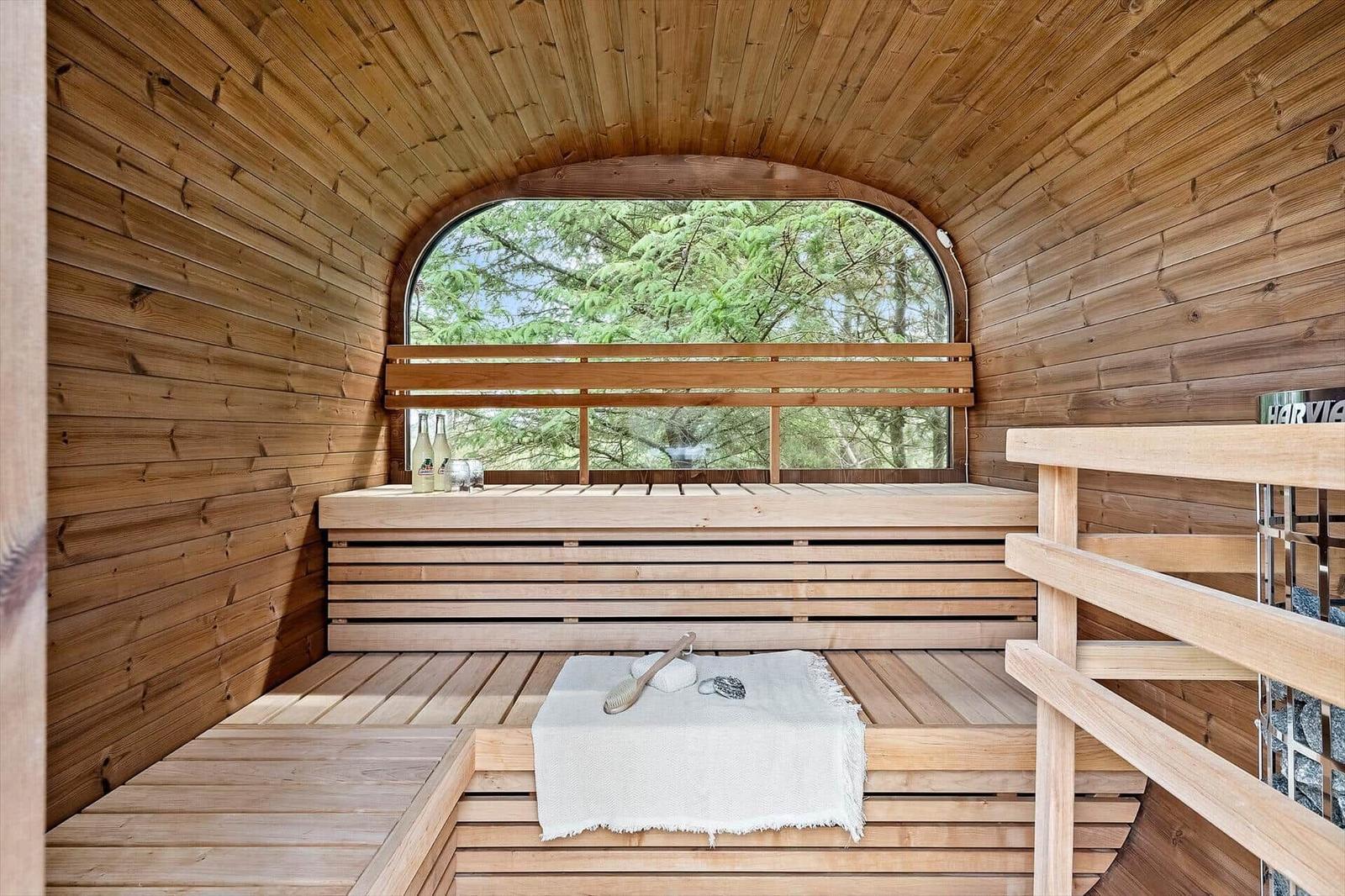 Holzsauna mit Sitzbänken und Bierflaschen vor großem Bogenfenster mit Waldblick.
