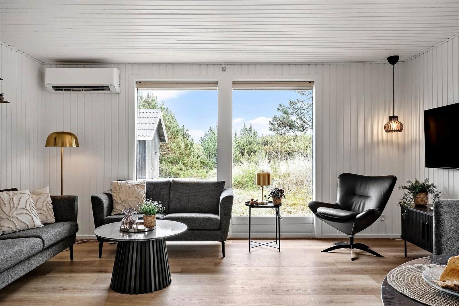 Wohnzimmer mit grauem Sofa, schwarzer Ledersessel und Holzboden. Große Fenster mit Blick auf Grünflächen.