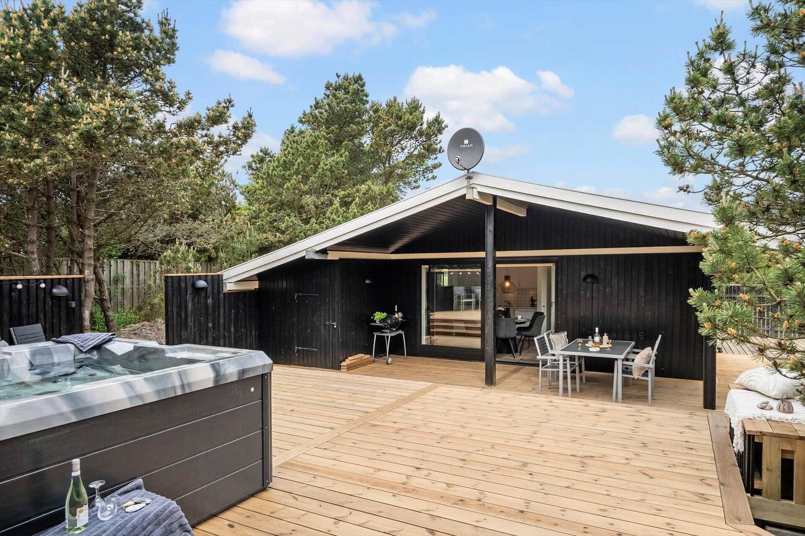 Moderne Holzhütte mit Terrasse, Whirlpool und Grillplatz unter Bäumen.