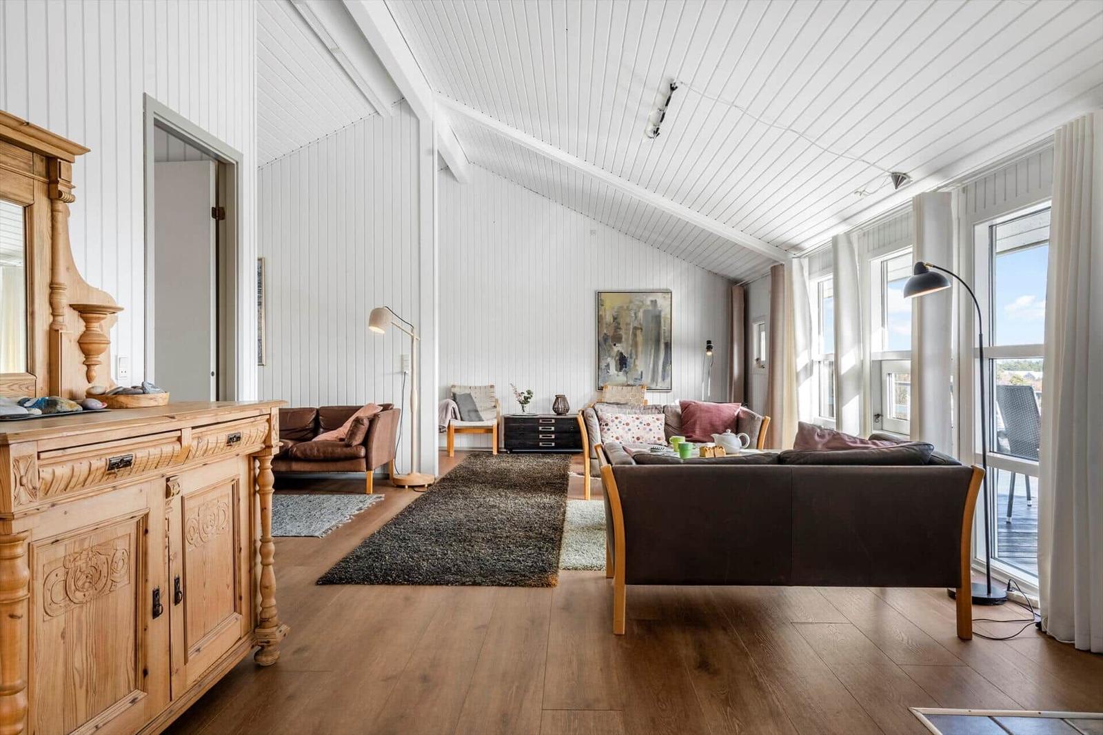 Wohnzimmer mit Holzboden, weißen Wänden und großem Fenster. Möbel und Teppich sind sichtbar.
