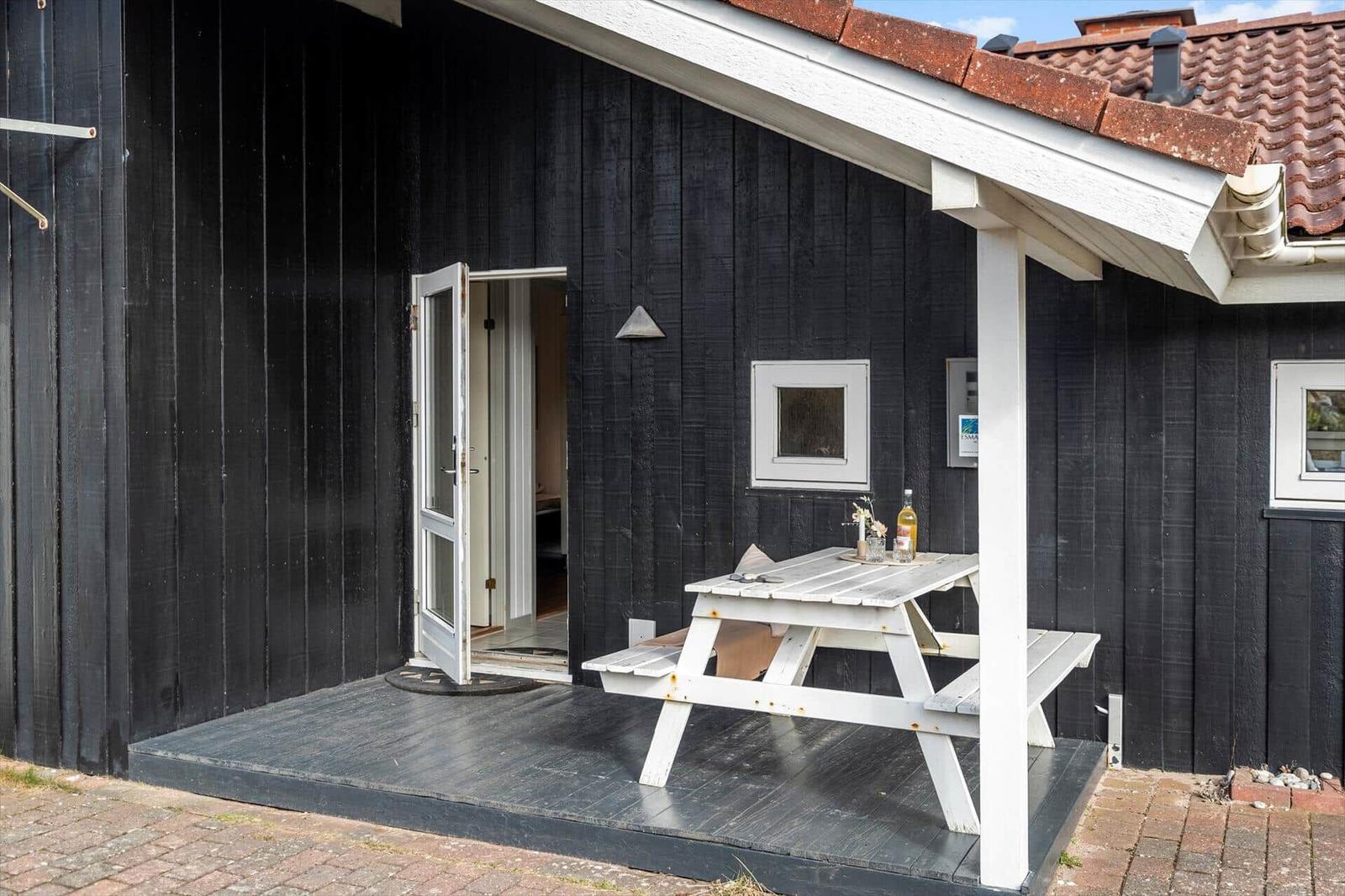 Helle Terrasse mit weißem Picknicktisch vor schwarzem Holzhaus mit offener Tür.