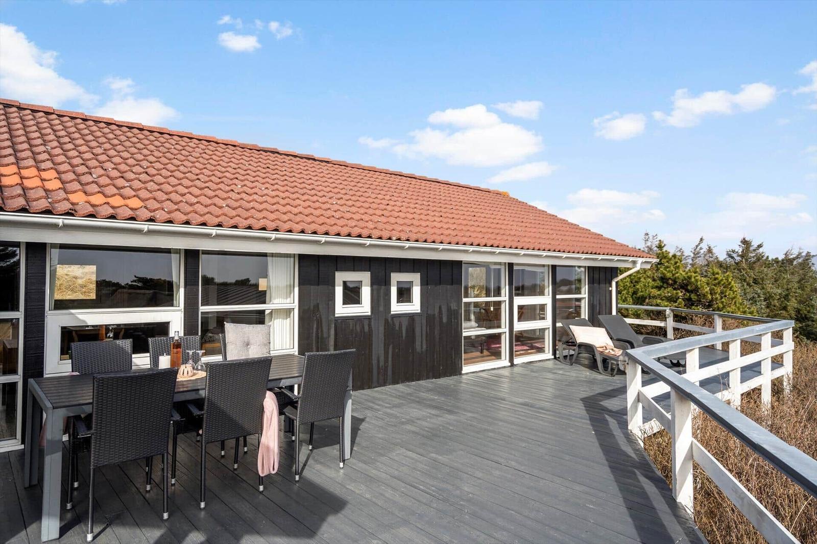 Terrasse mit Tisch, Stühlen und Liegen. Haus mit rotem Dach und dunkler Fassade.