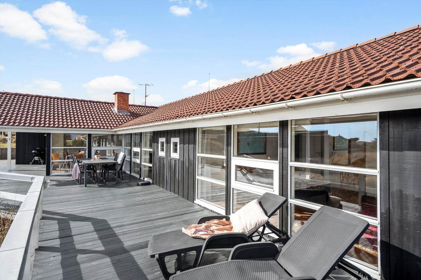Terrasse mit Liegestühlen und Tisch. Haus mit rotem Dach und großen Fenstern.