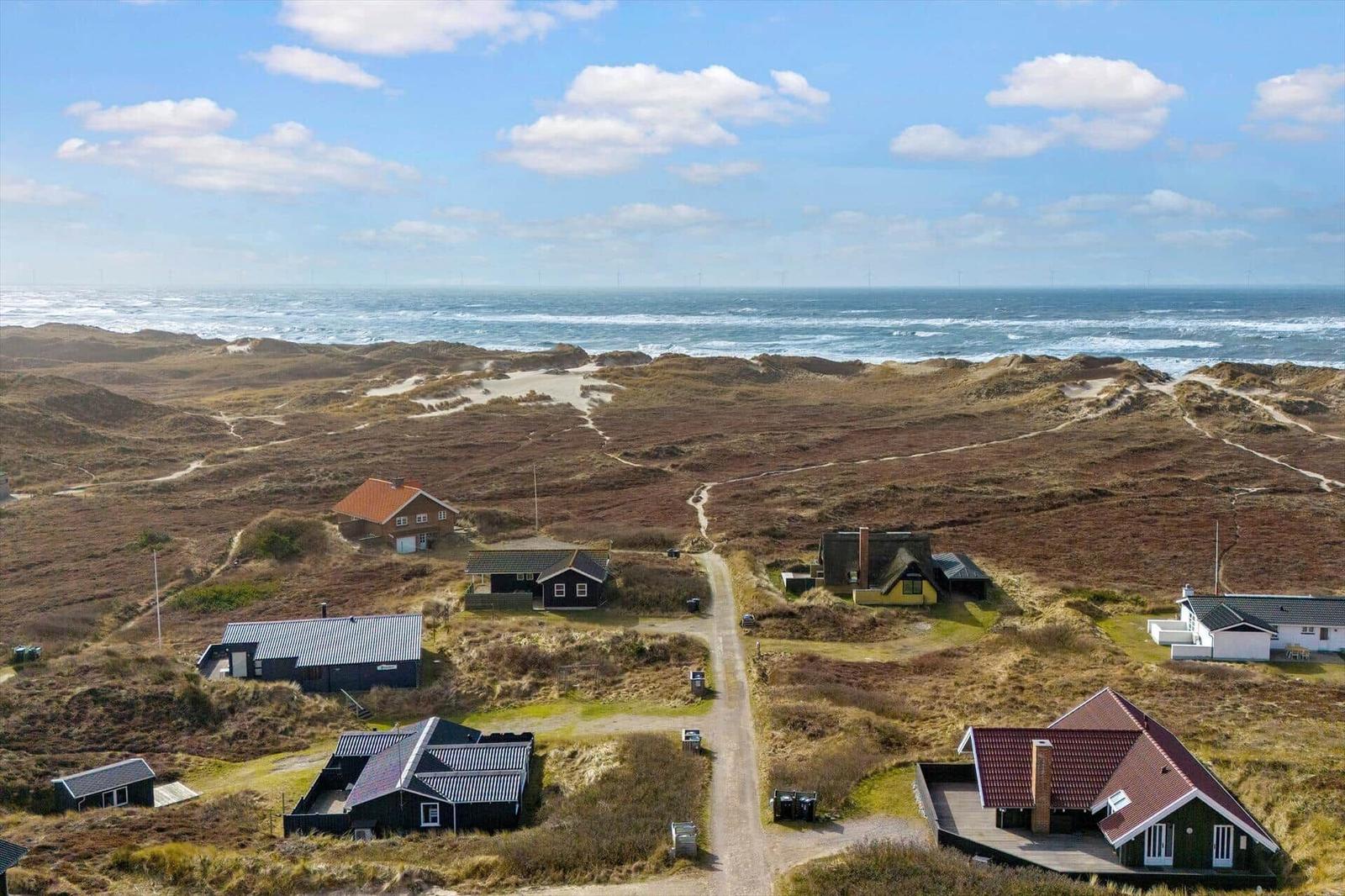 Strandnahe Häuser auf Dünen mit Blick auf das Meer und Windkraftanlagen im Hintergrund.