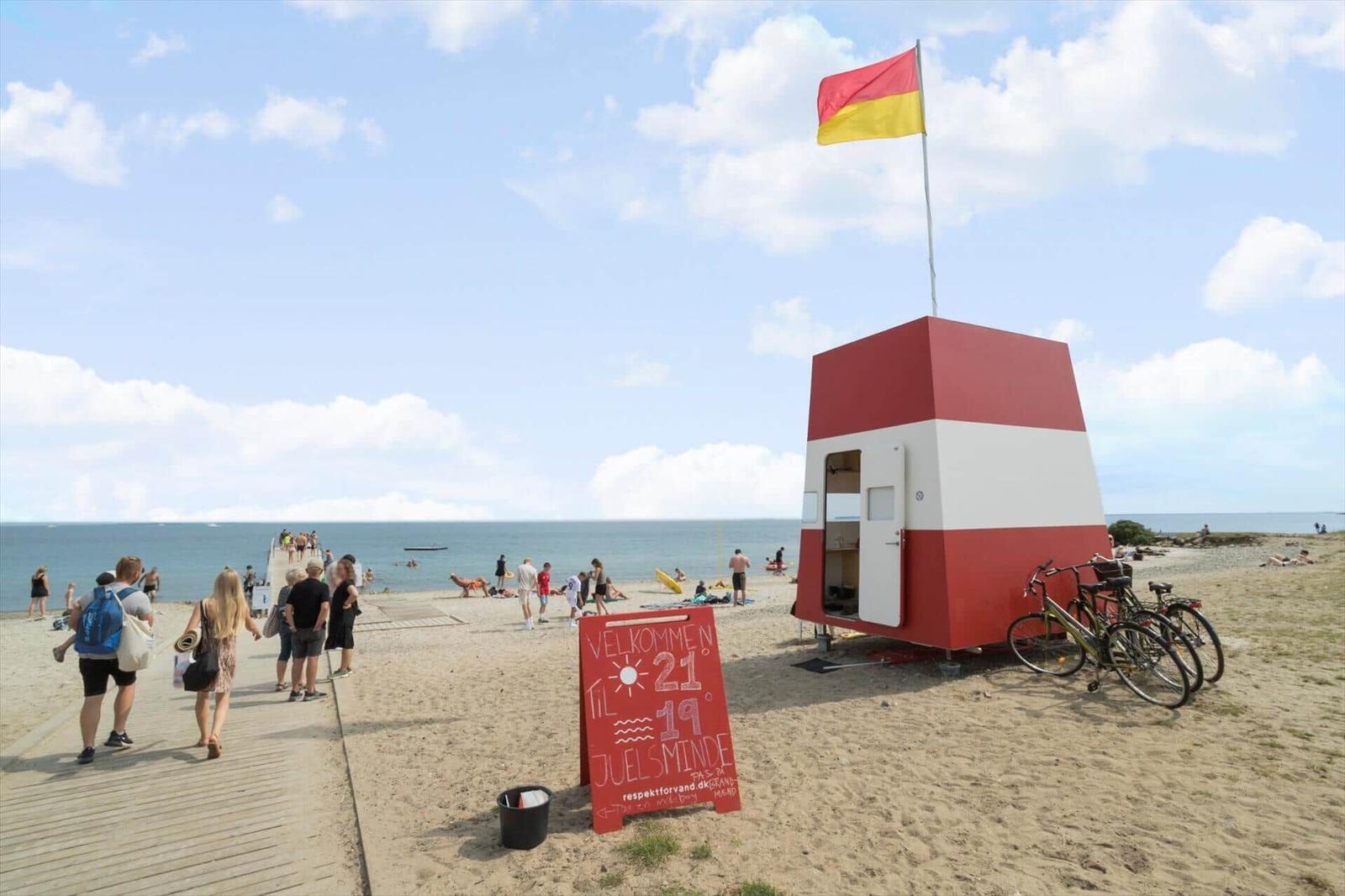Beach with lifeguard station, flag, and sign. People stroll and swim.