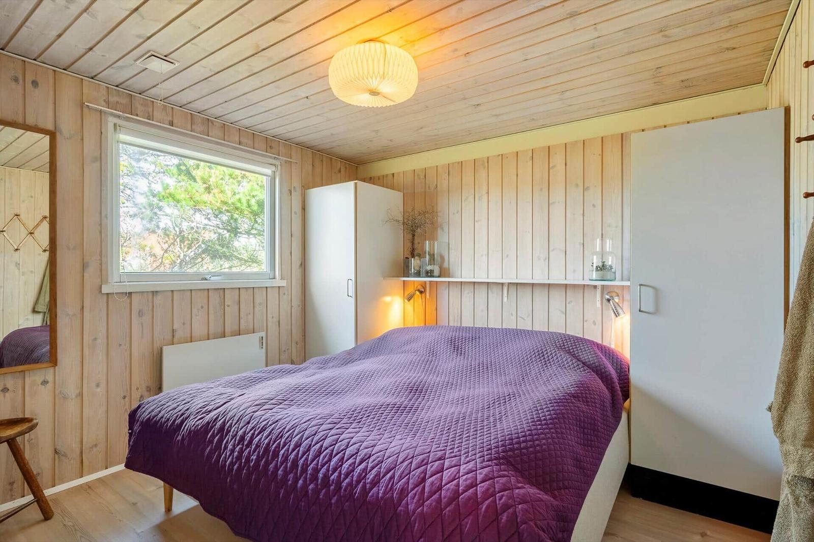 Schlafzimmer mit Holzwand, Bett mit lila Daunendecke und Fenster mit Blick auf Bäume.