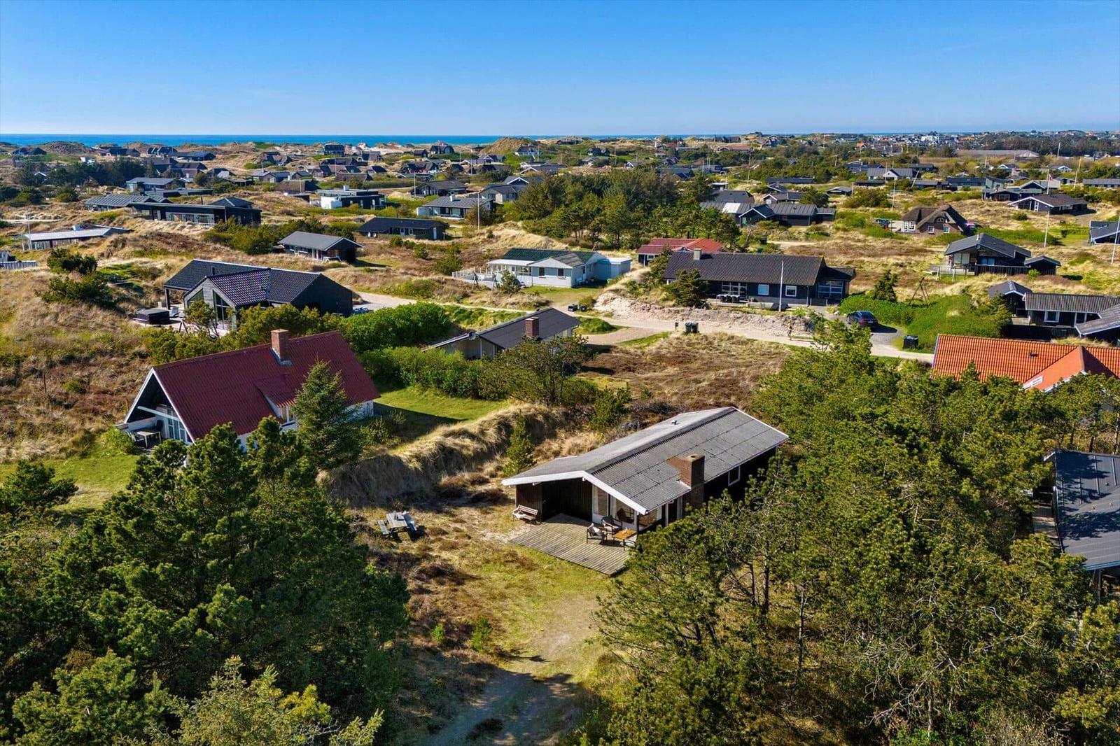 Haus mit Terrasse zwischen Bäumen, Blick auf Strand und Dünenlandschaft.
