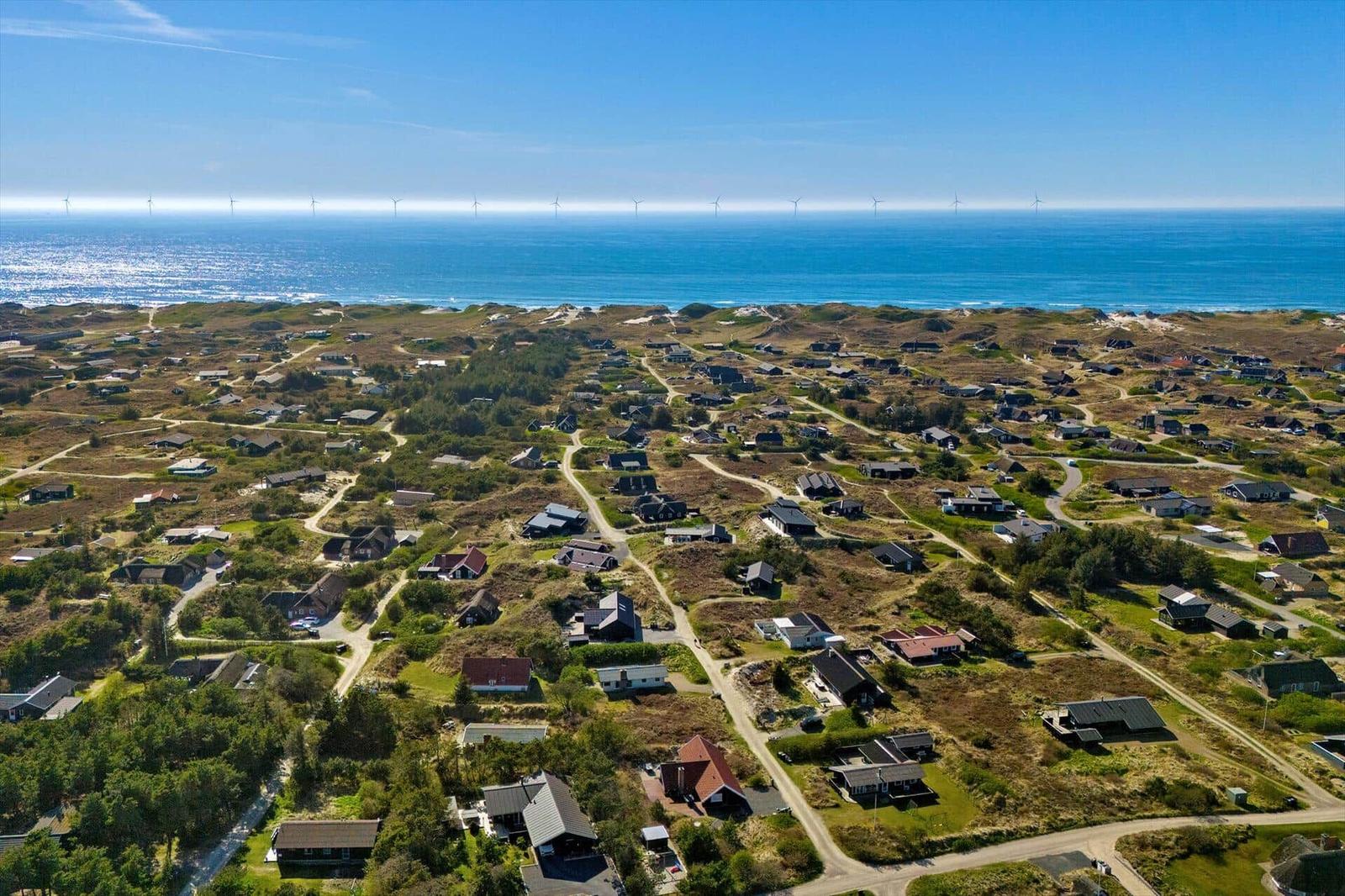 Aerial view of coastal village with houses, dunes, and wind turbines on horizon.