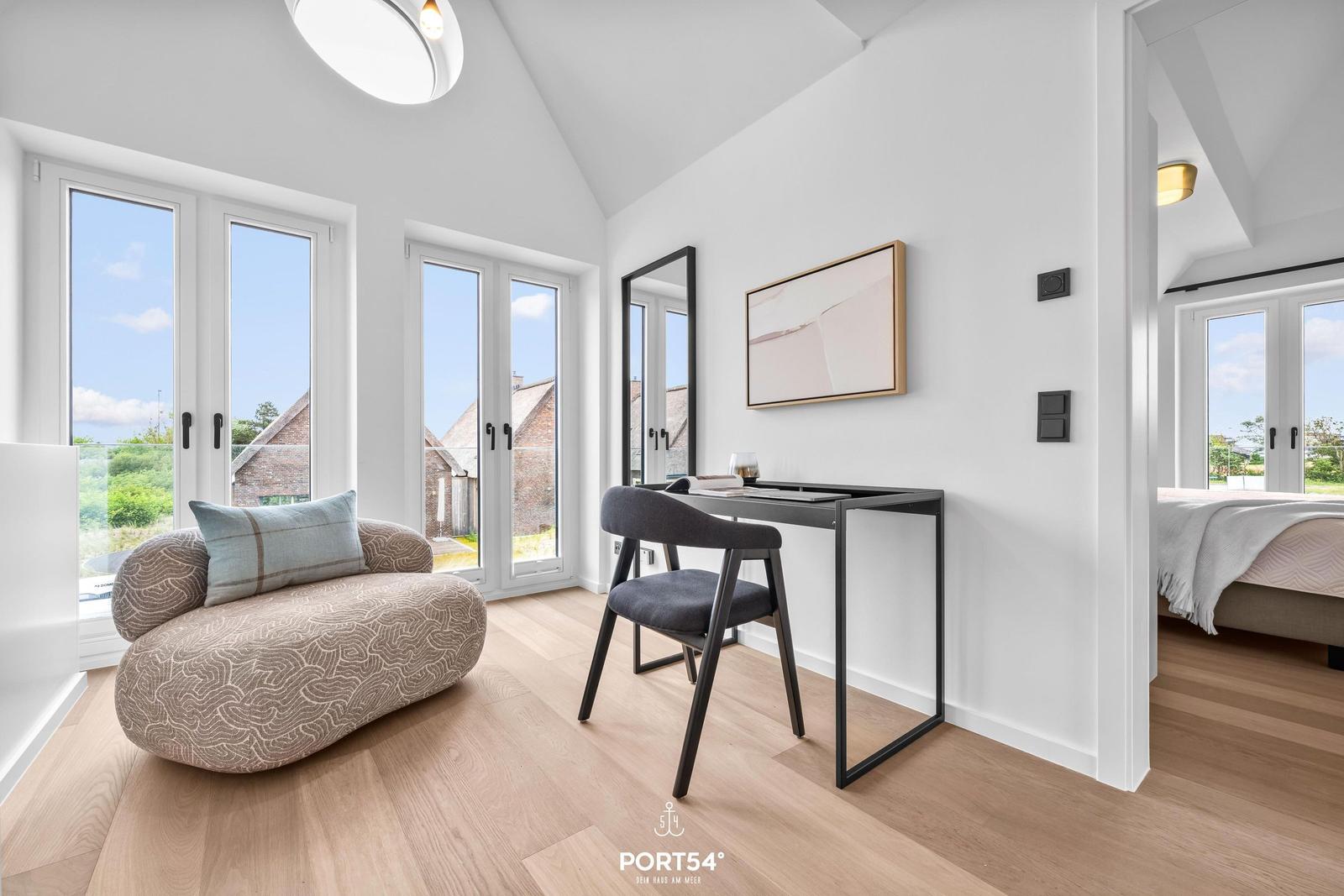 Ein Raum mit Schreibtisch, Sessel und großen Fenstern. Man sieht einen Blick in ein Schlafzimmer.