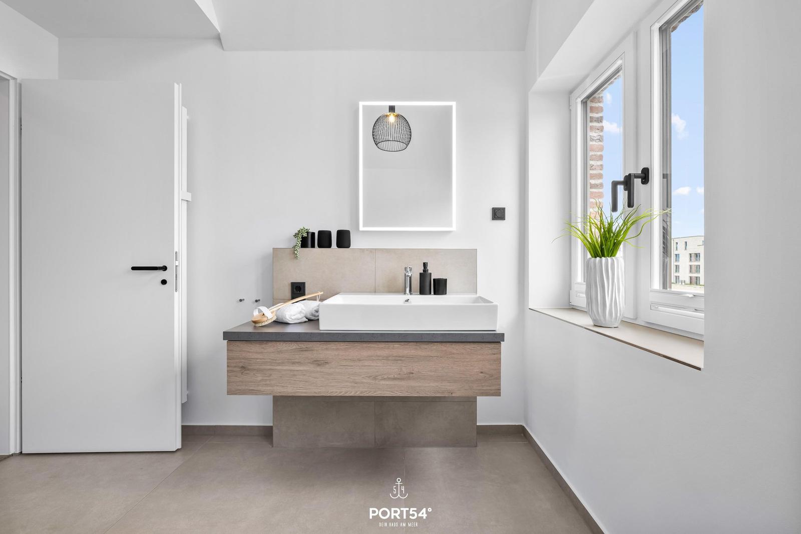 Badezimmer mit Waschtisch, Spiegel und Fenster mit Pflanze.