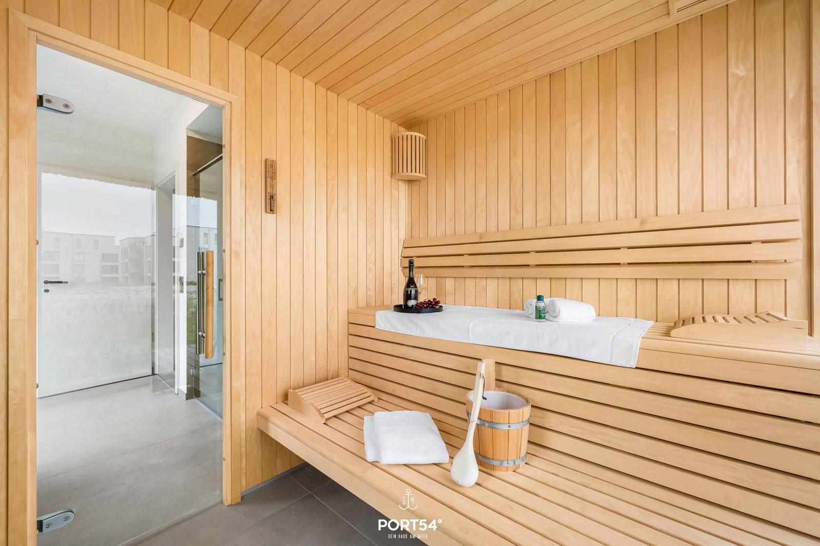 Sauna mit Holzbänken, Handtuch, Eimer und Flasche. Tür führt zu einem Bad.