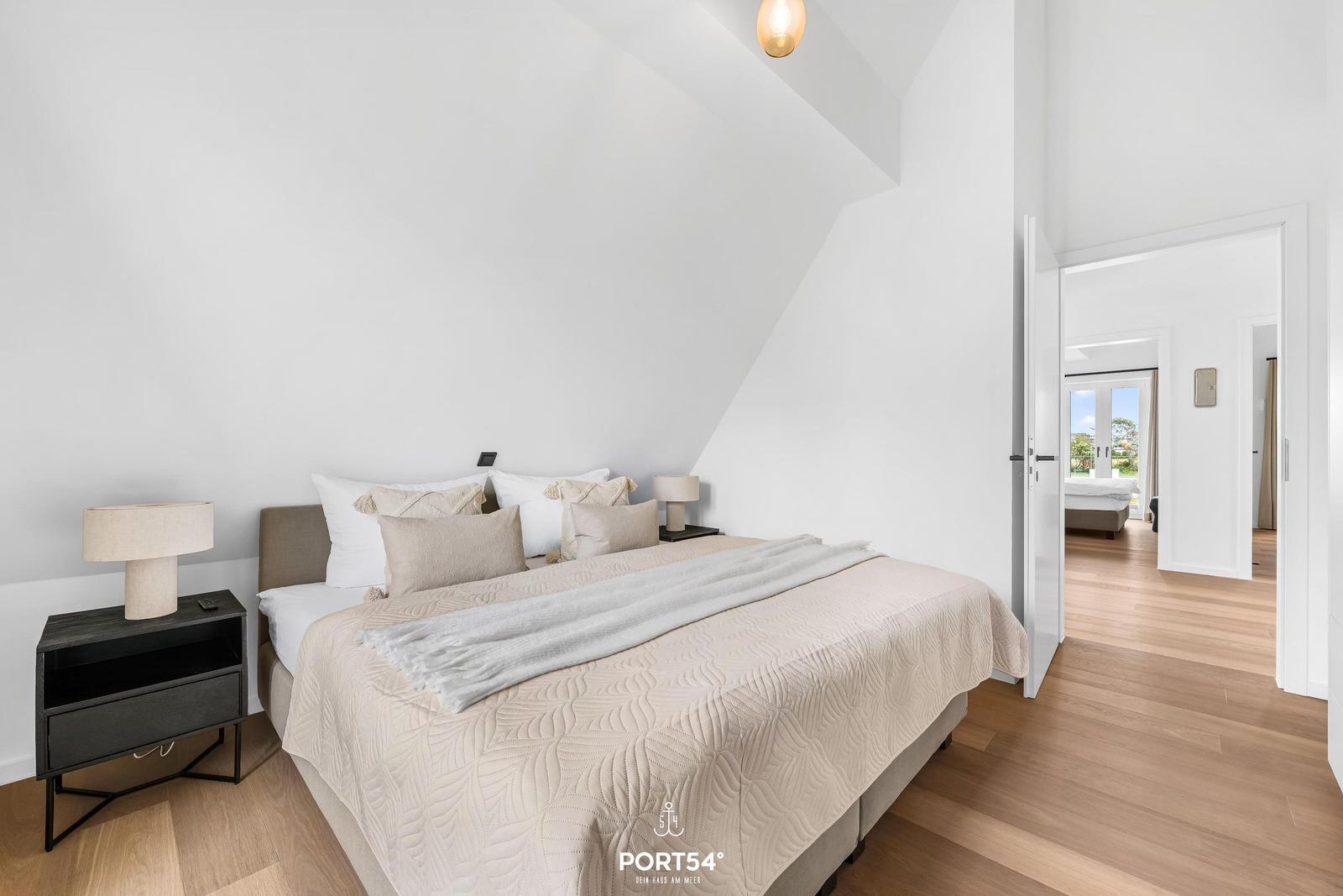 Schlafzimmer mit Bett, Nachttischen und Holzboden. Tür führt in einen weiteren Raum.