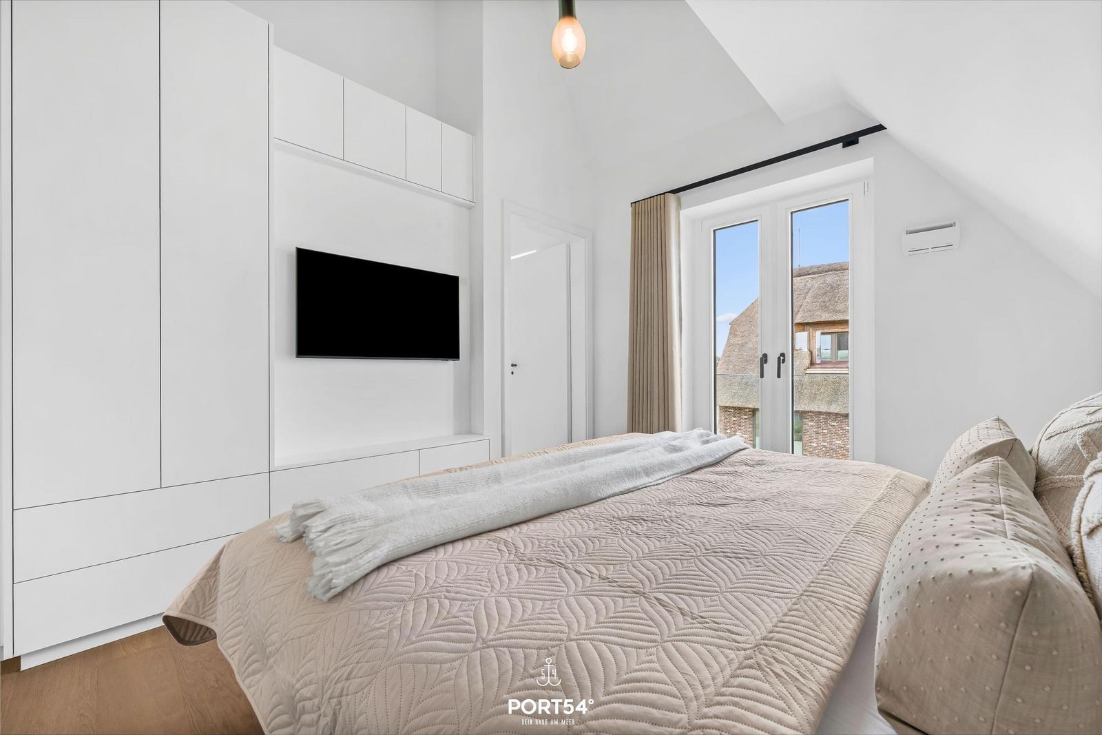 Schlafzimmer mit Bett, TV und Fenster mit Blick auf Dächer.