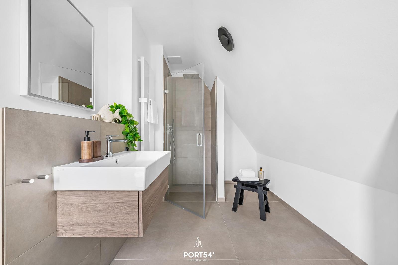 Badezimmer mit Dusche, Waschbecken und Spiegel. Dachneigung sichtbar.