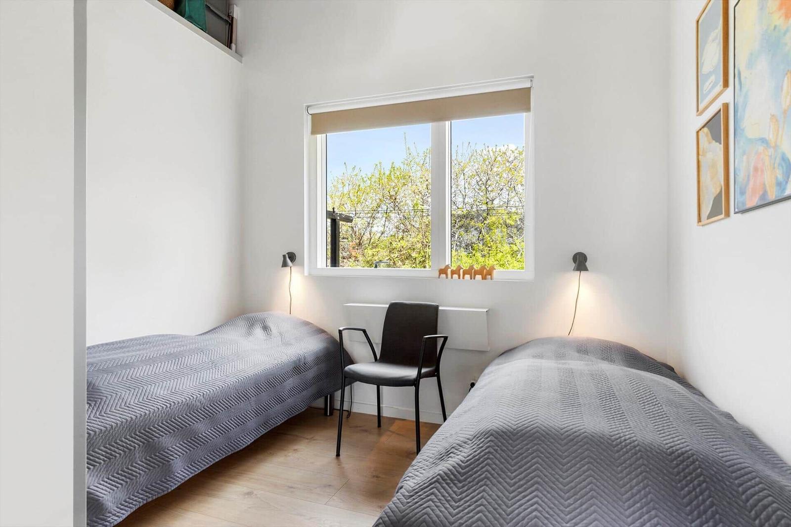 Zwei Betten mit grauer Bettwäsche, ein Fenster mit Blick auf Bäume und eine schwarze Stuhl