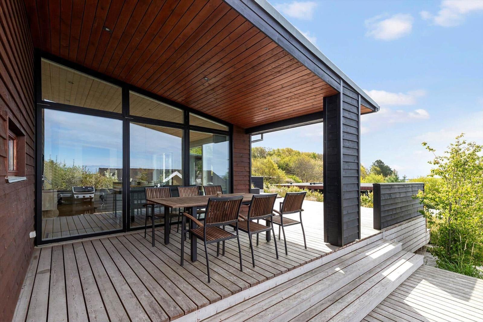 Terrasse mit Tisch und Stühlen unter Holzüberdachung. Glaswände und Blick auf Grünflächen.