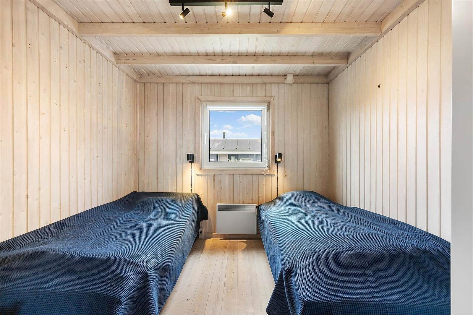 Zwei Betten mit blauen Bettdecken in einem holzverkleideten Raum mit Fenster und Heizung.
