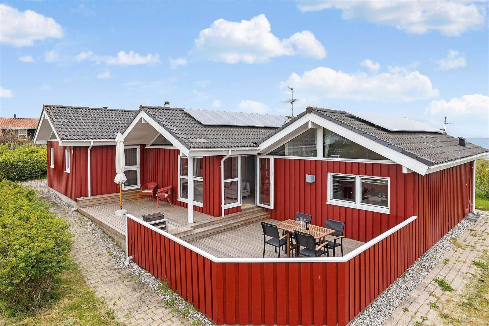 Rotes Holzhaus mit Terrasse, Garten und Solaranlage unter blauem Himmel.