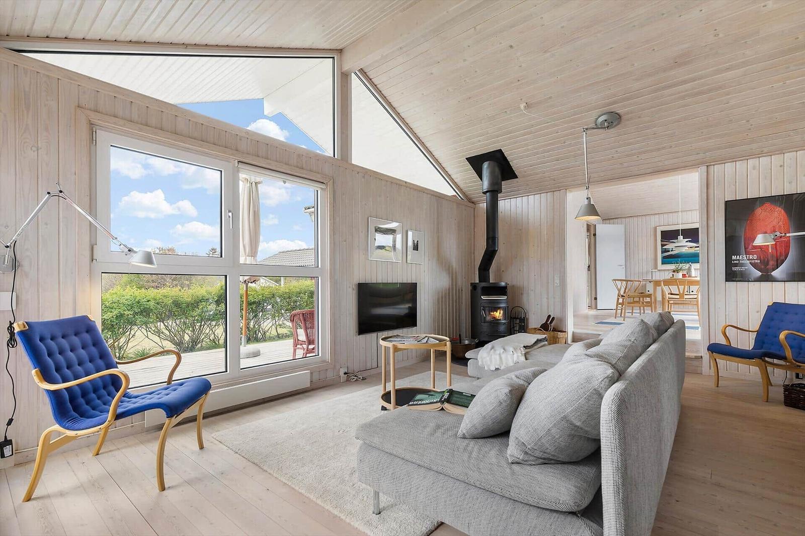 Wohnzimmer mit Sofa, Kamin und großen Fenstern. Blaue Sessel und Holzwände.