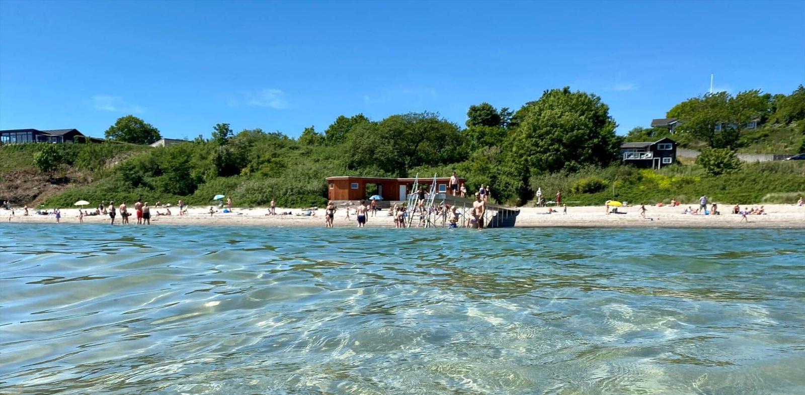 Beach with bathers, wooden diving platform, and green hills in background.